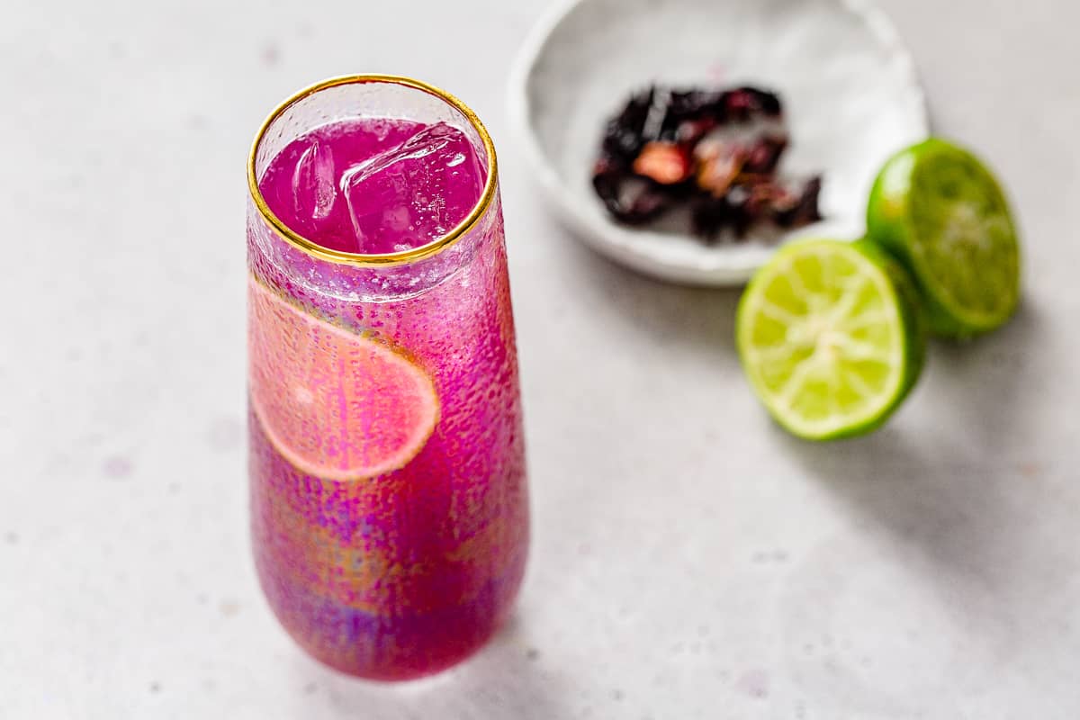 View of the top of a hibiscus gin cocktail with dried hibiscus flowers and fresh lime in the background.