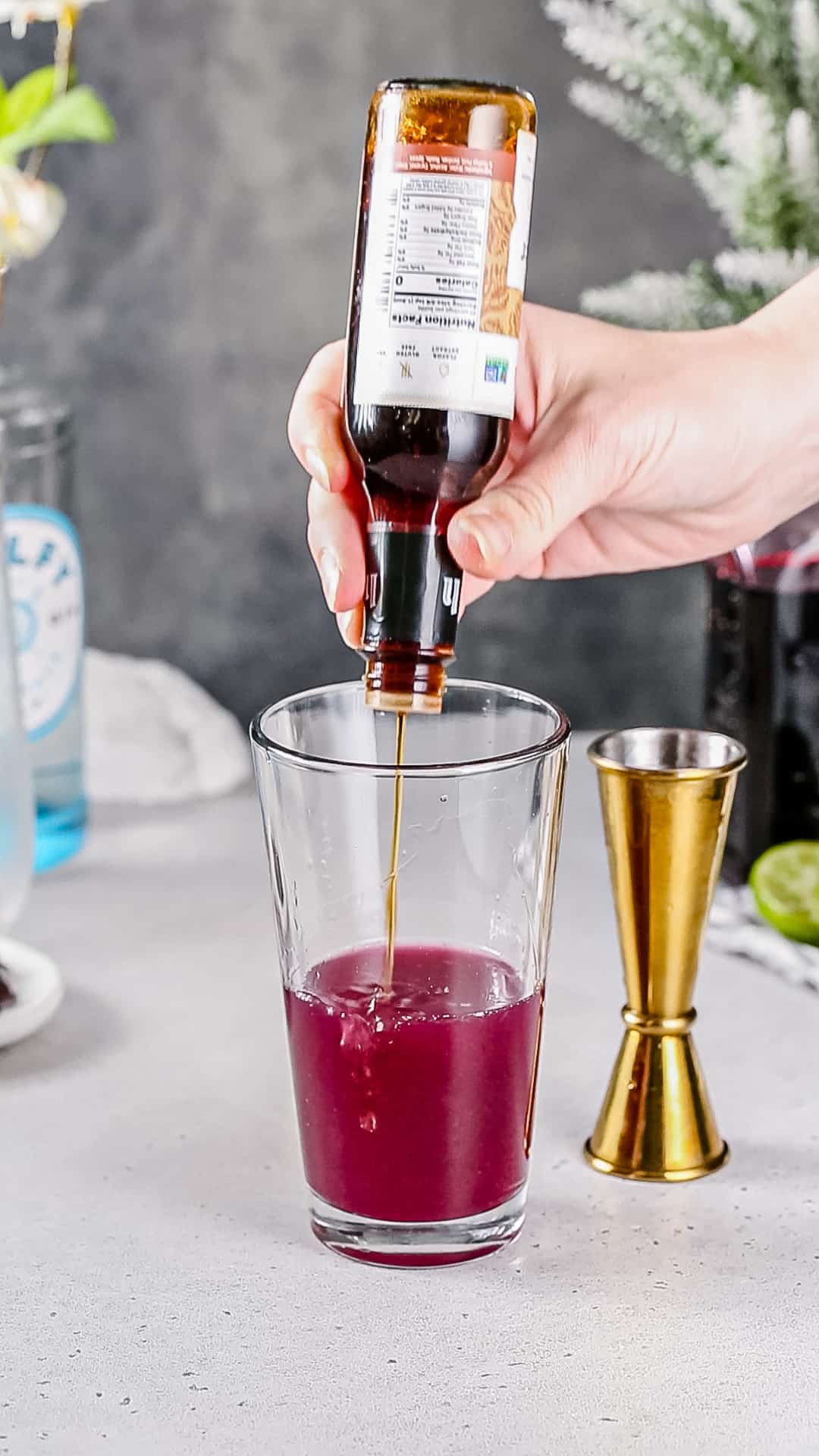 Adding ginger bitters to a cocktail shaker filled with dark pink liquid.