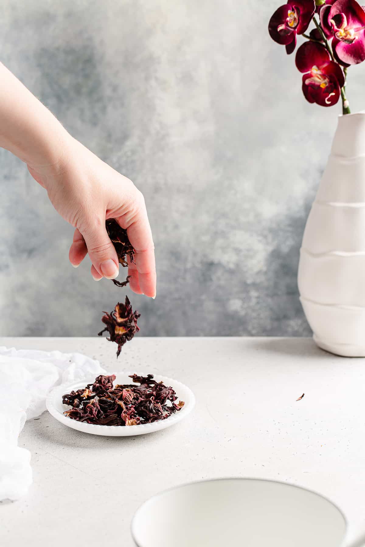 Hand dropping dried hibiscus flowers onto a plate with purple orchids in a vase in the background.