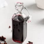 Open bottle of hibiscus syrup on a countertop with dried hibiscus flowers on the side.