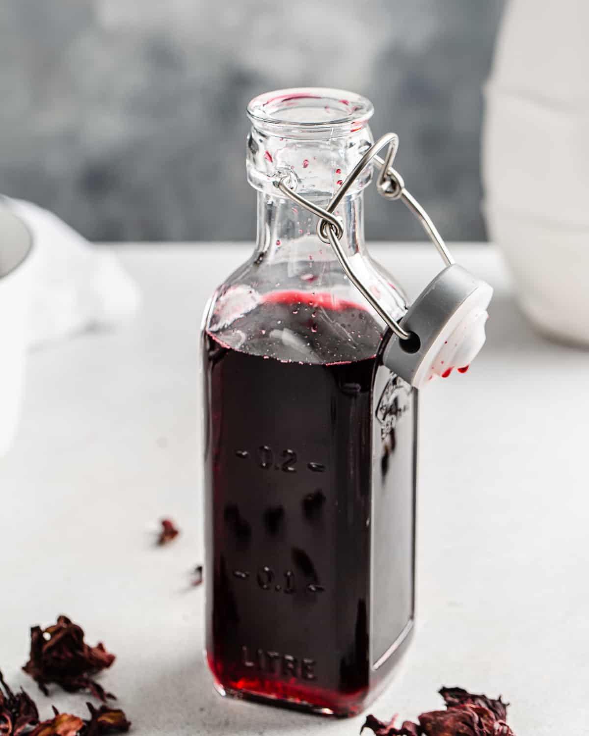 Close up of a bottle of hibiscus syrup with dried hibiscus flowers around it.
