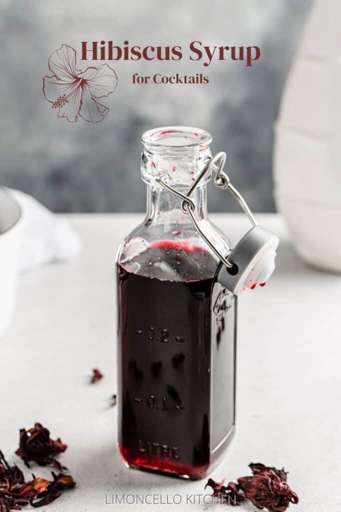 Bottle of hibiscus syrup on a countertop with dried hibiscus flowers around it and the text Hibiscus Syrup for Cocktails on top.