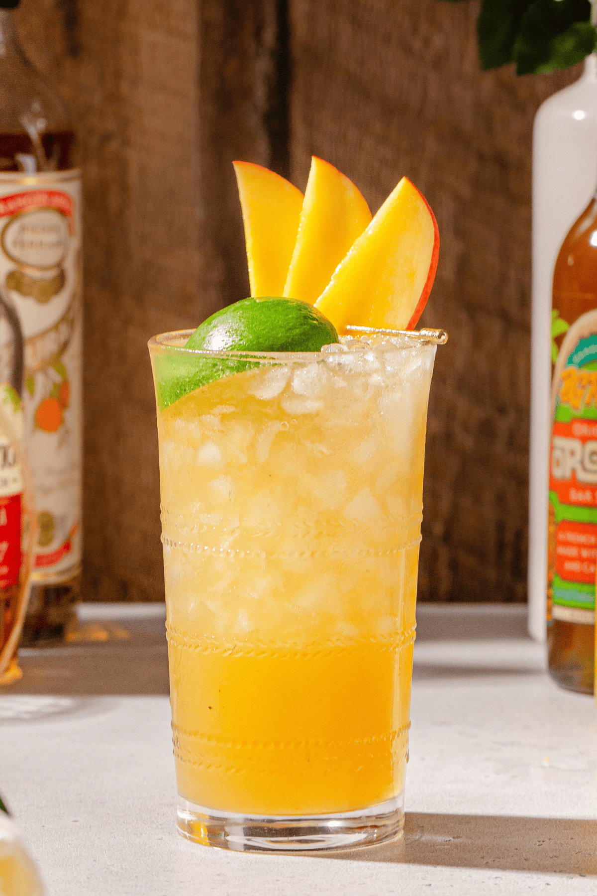 Mango Mai Tai on a countertop garnished with mango slices and a lime. Some bottles used to make the drink are seen in the background.