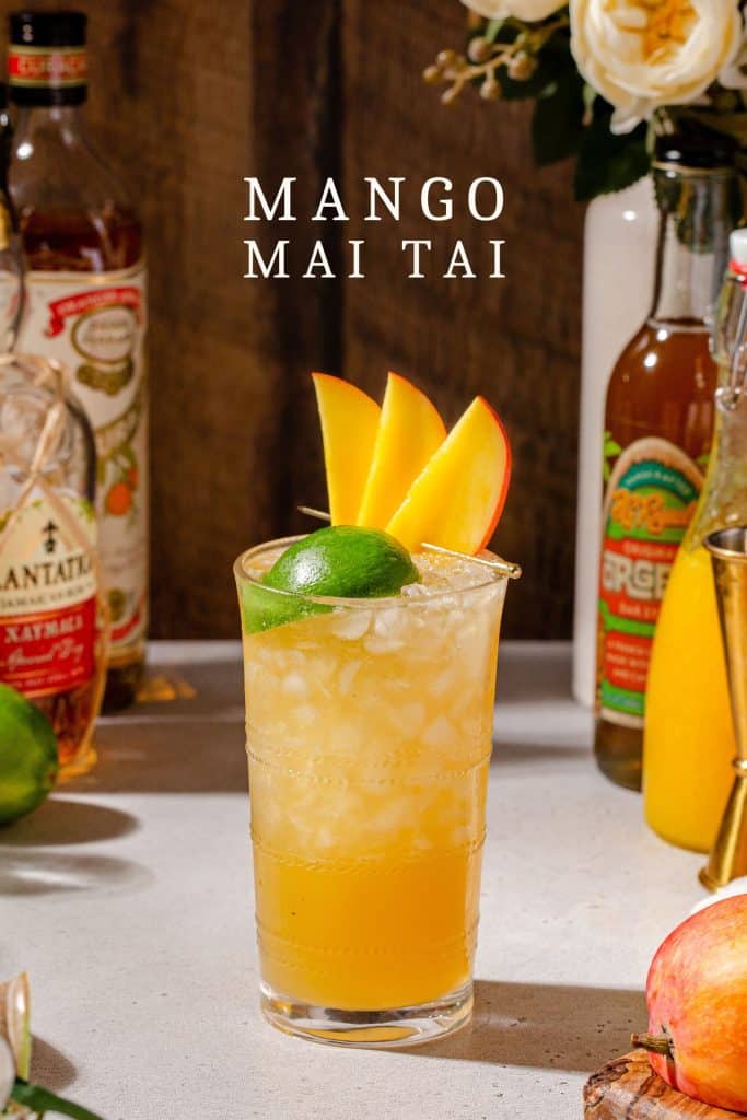 Side view of a Mango mai tai cocktail on a countertop surrounded by ingredients used to make the drink. Text above the drink says "mango mai tai".