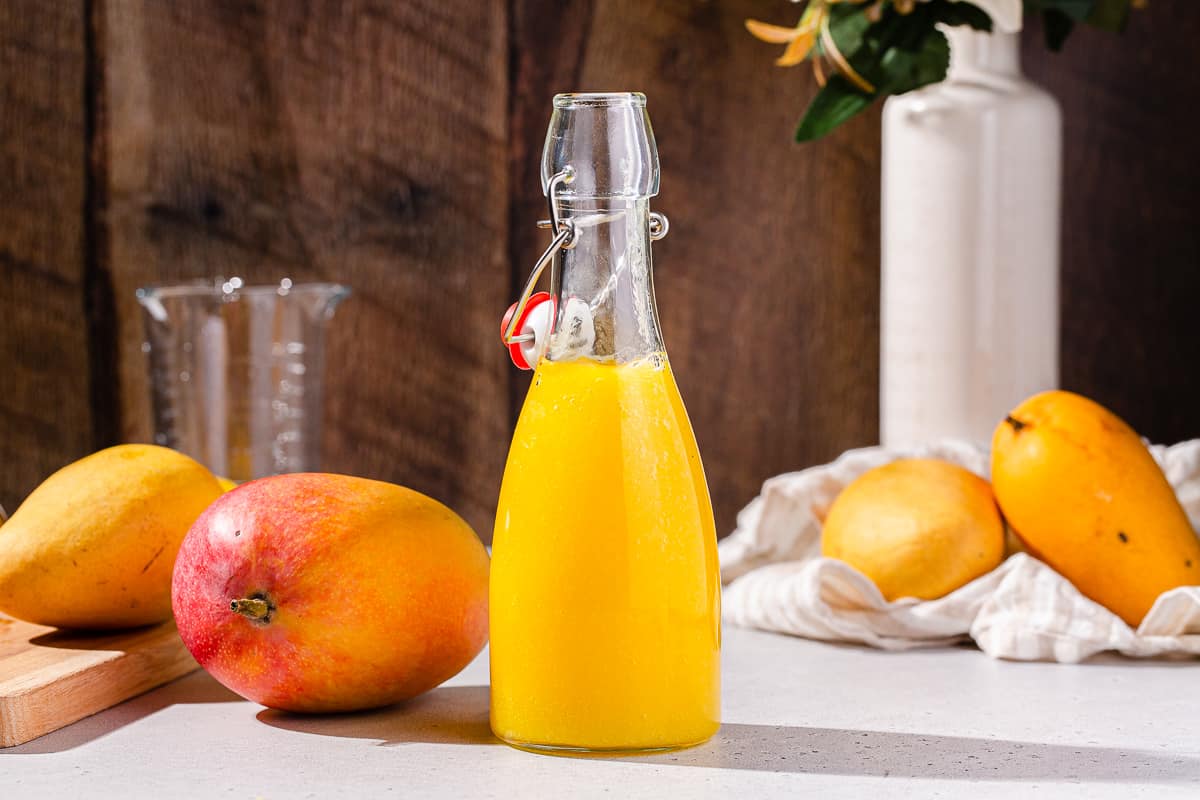 Side view of mango nectar in a glass bottle with mangoes around it and flowers in the background.