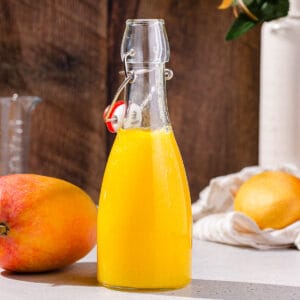 Side view of mango nectar in a glass bottle with mangoes around it and flowers in the background.