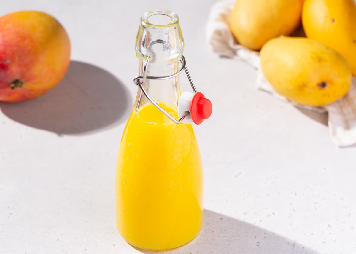Mango nectar in a glass bottle with two types of mangoes in the background.