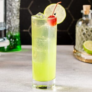 Side view of a Tokyo Tea cocktail, which is chartreuse green in color and has a lime wheel garnish on the rim and a maraschino cherry next to the lime. The cocktail is in a tall glass with straight sides and has ice cubes in it. In the background are a bottle of Midori melon along with a bottle of gin, a lime, and a bottle of simple syrup.