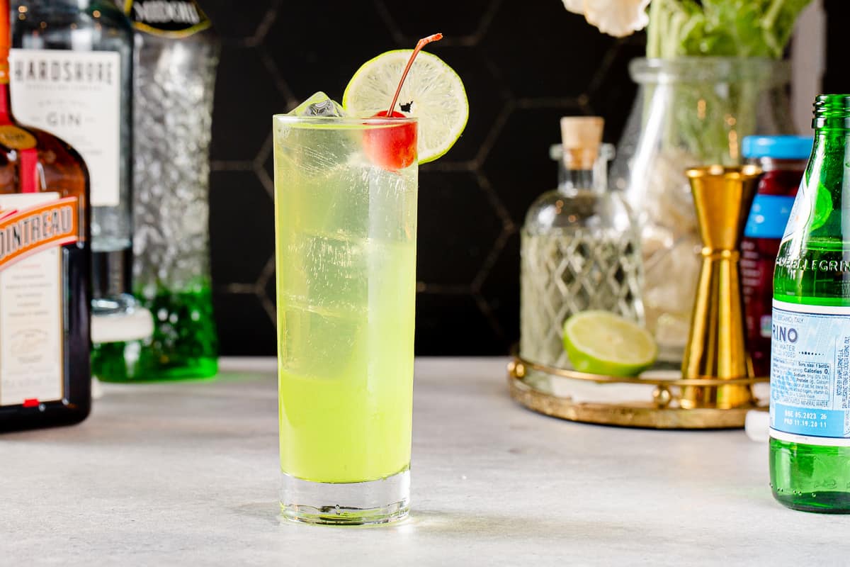 Side view of a Tokyo Tea cocktail, which is chartreuse green in color and has a lime wheel garnish on the rim and a maraschino cherry next to the lime. The cocktail is in a tall glass with straight sides and has ice cubes in it. In the background are various ingredients along with a vase of flowers and bar tools..