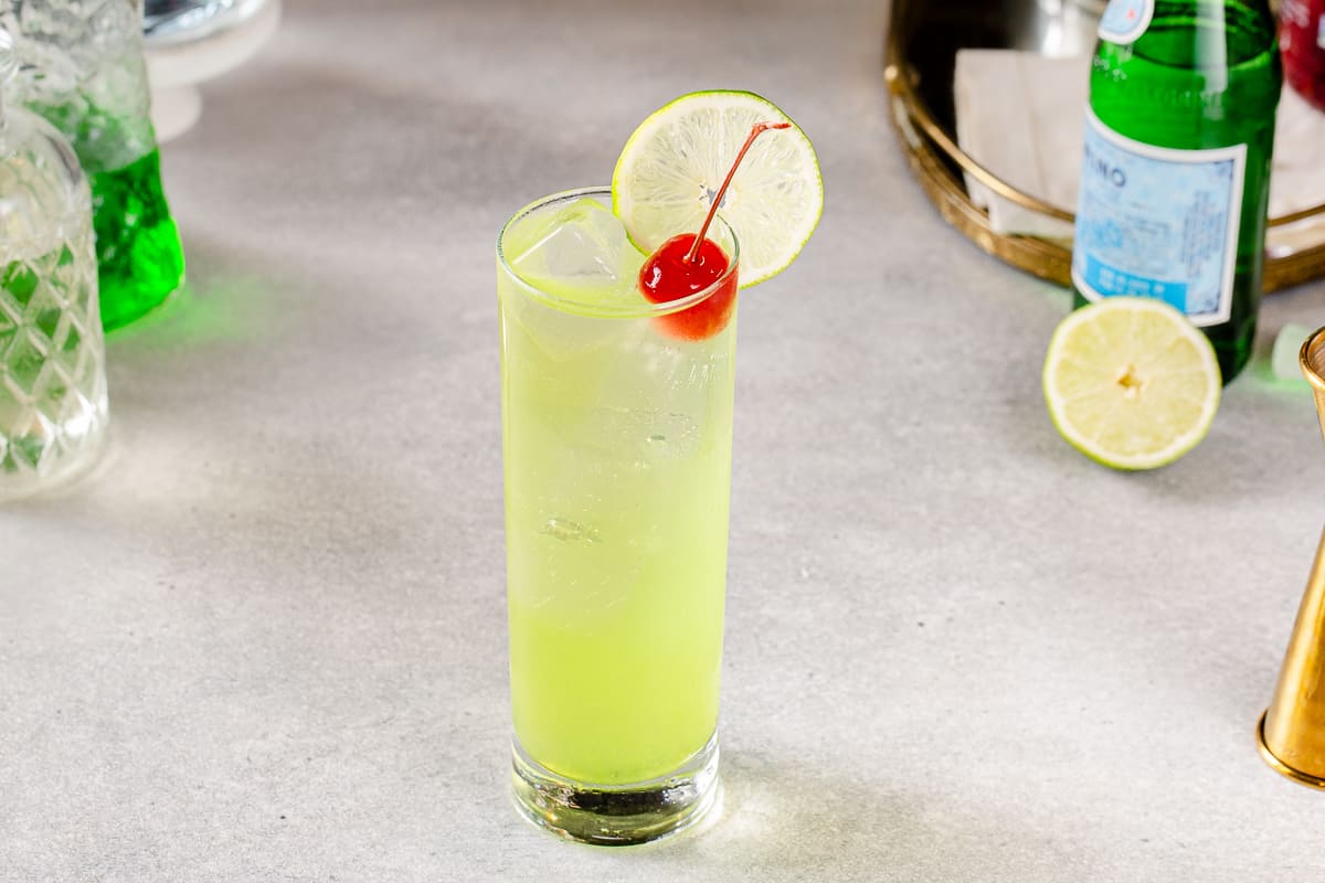 Semi-overhead view of a Tokyo Tea cocktail in a tall Collins glass, which has straight sides. The drink is chartreuse green and has a lime wheel and maraschino cherry garnish. Ingredients and a mirrored tray are in the background.