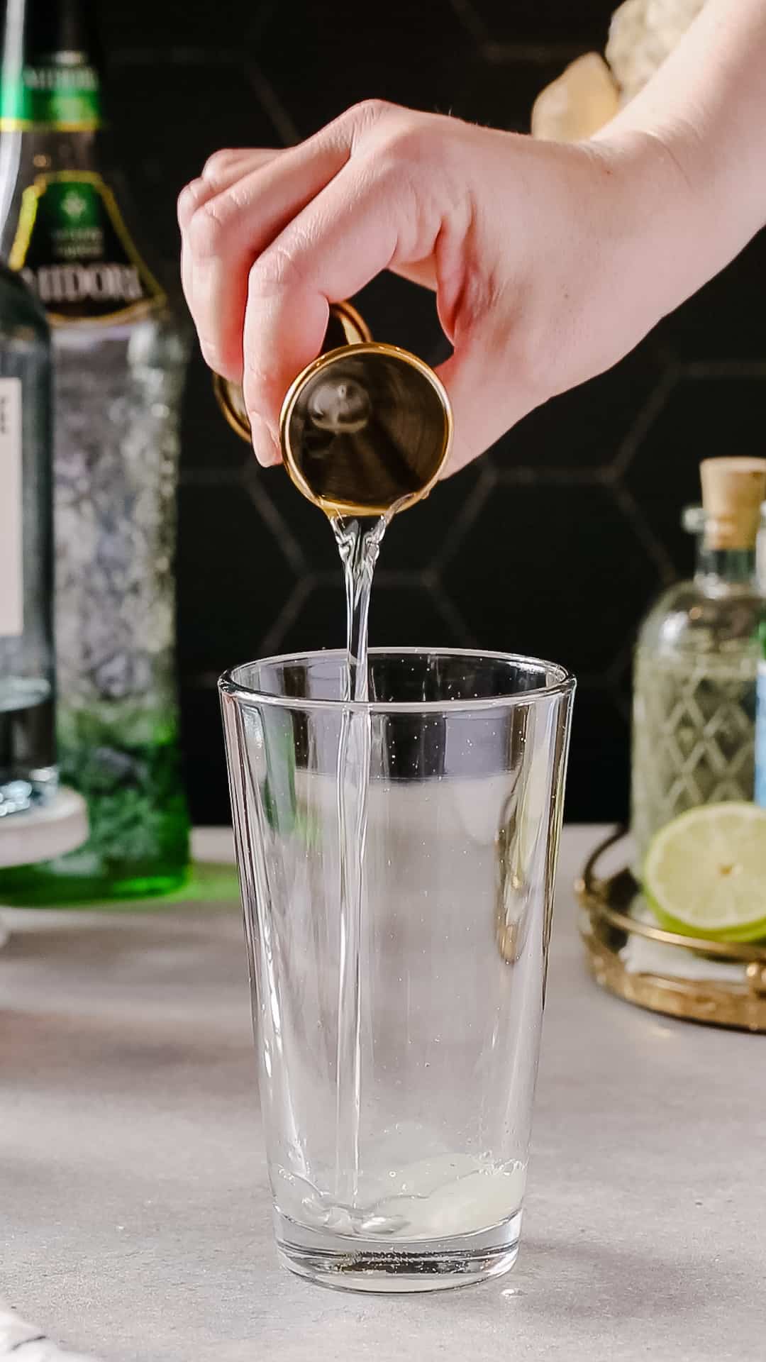 Hand pouring simple syrup from a jigger into a glass cocktail shaker.