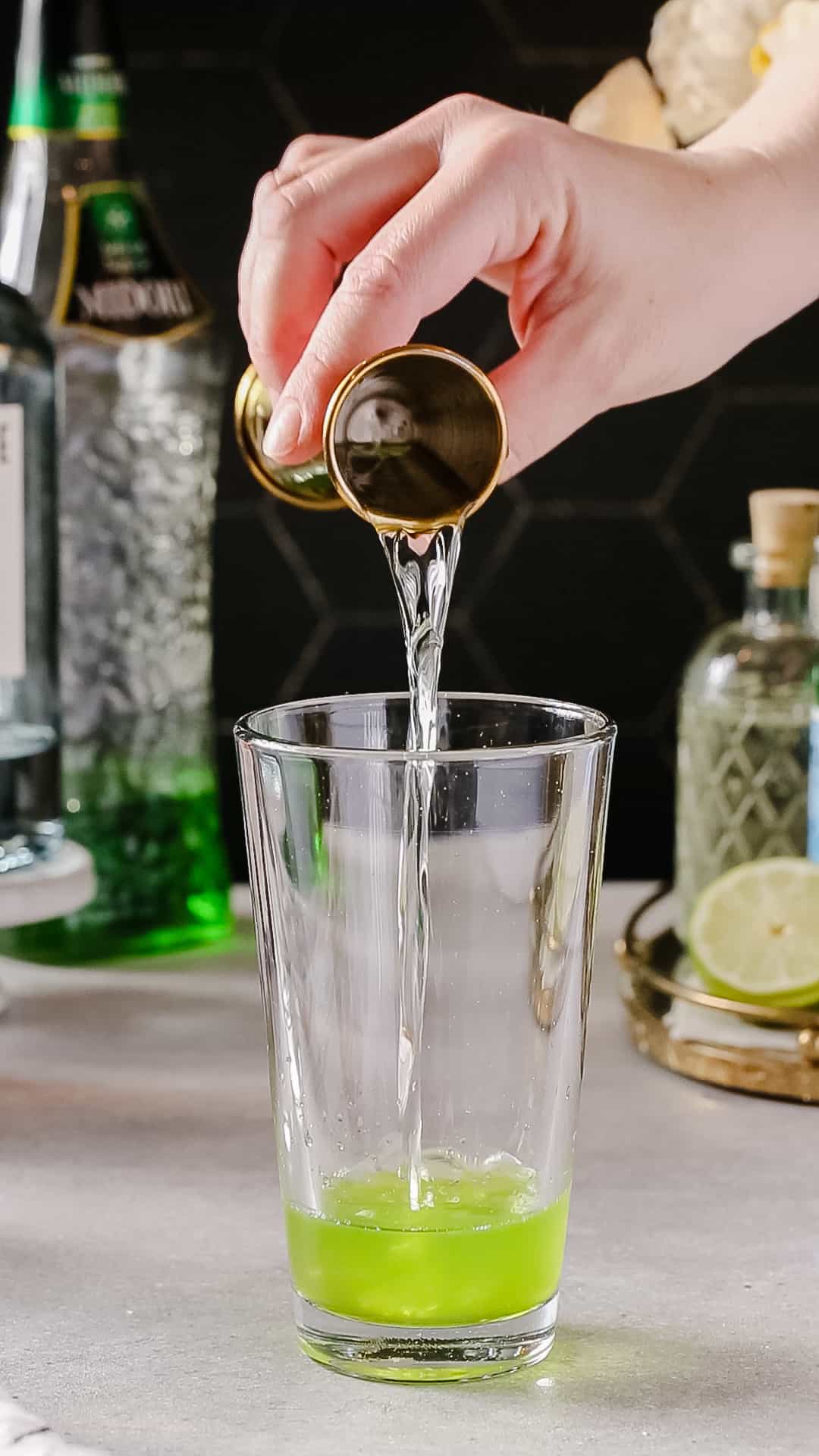 Hand pouring vodka from a jigger into a glass cocktail shaker.