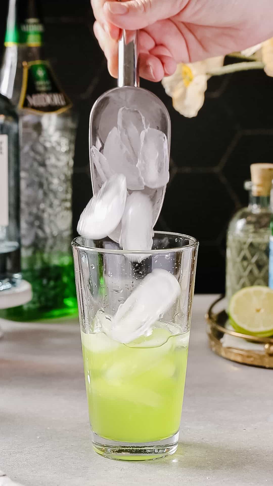 Hand using an ice scoop to add ice to a glass cocktail shaker filled with green liquid.
