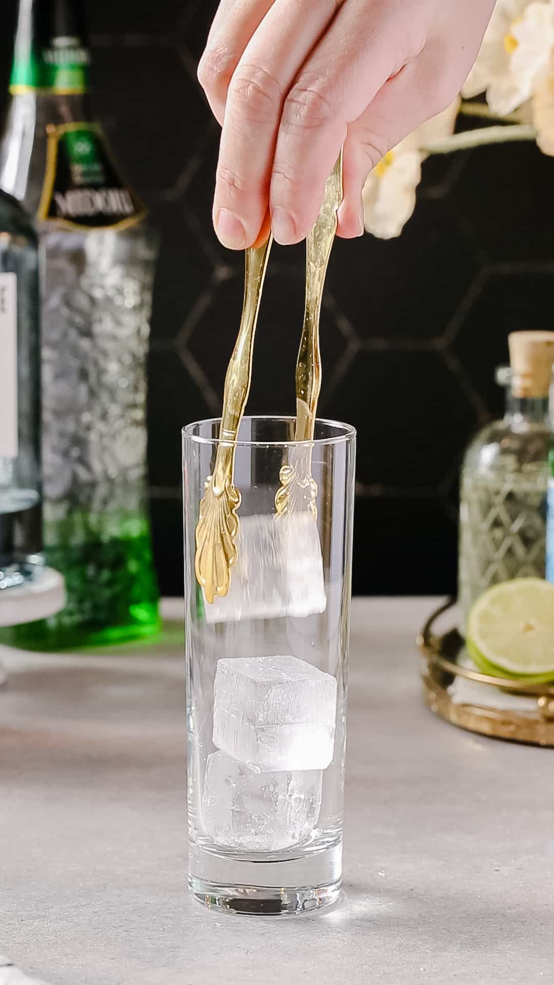Hands using gold colored ice tongs to add ice to a Collins cocktail glass.