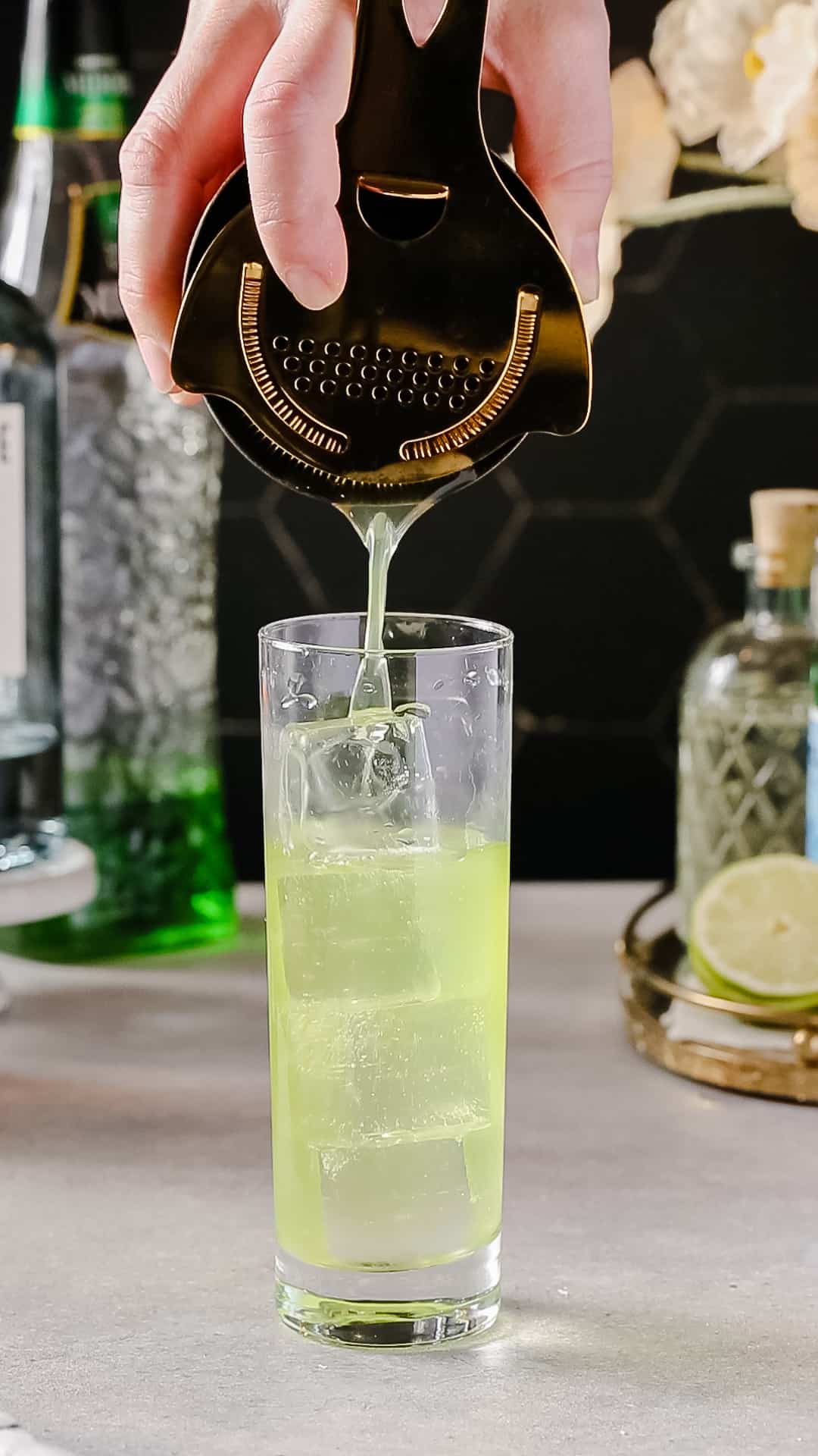 Hand using a gold colored cocktail shaker and strainer to strain a green colored drink into a Collins glass filled with ice.