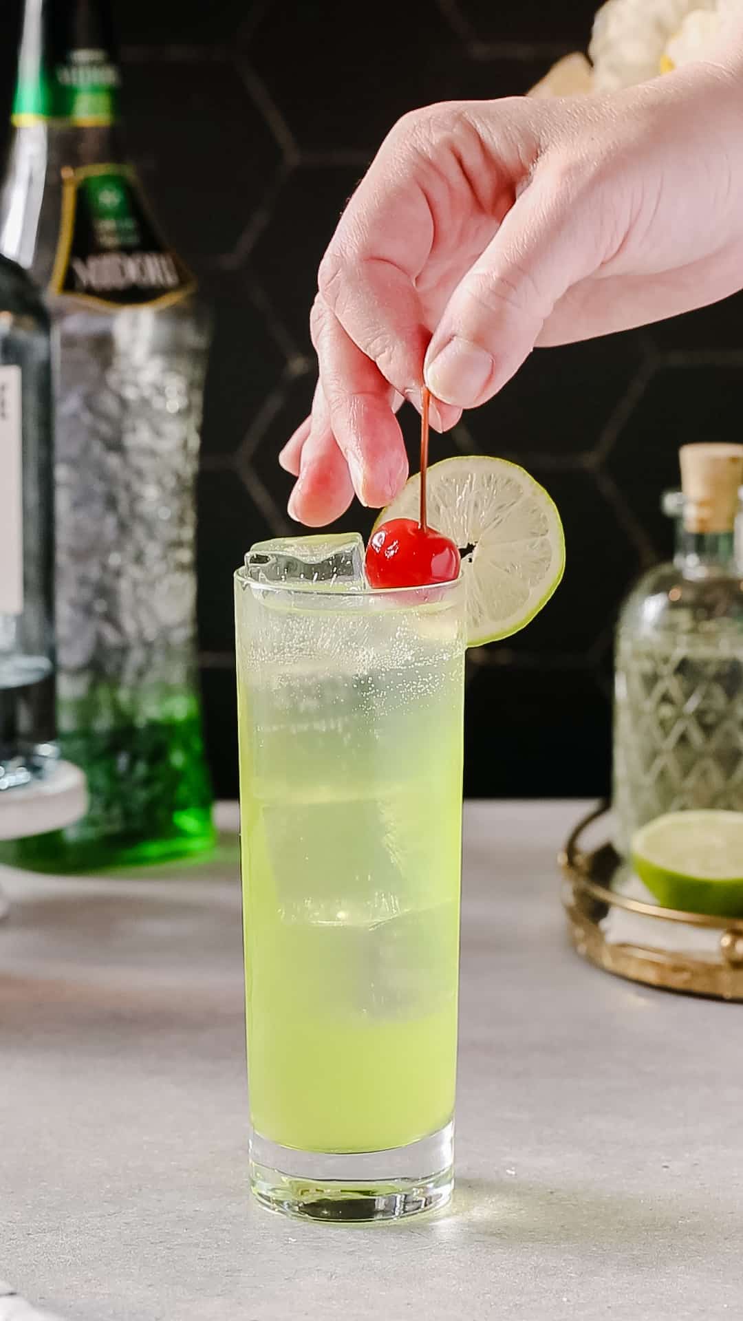 Hand adding a maraschino cherry to the top of a cocktail glass filled with green liquid and ice cubes.
