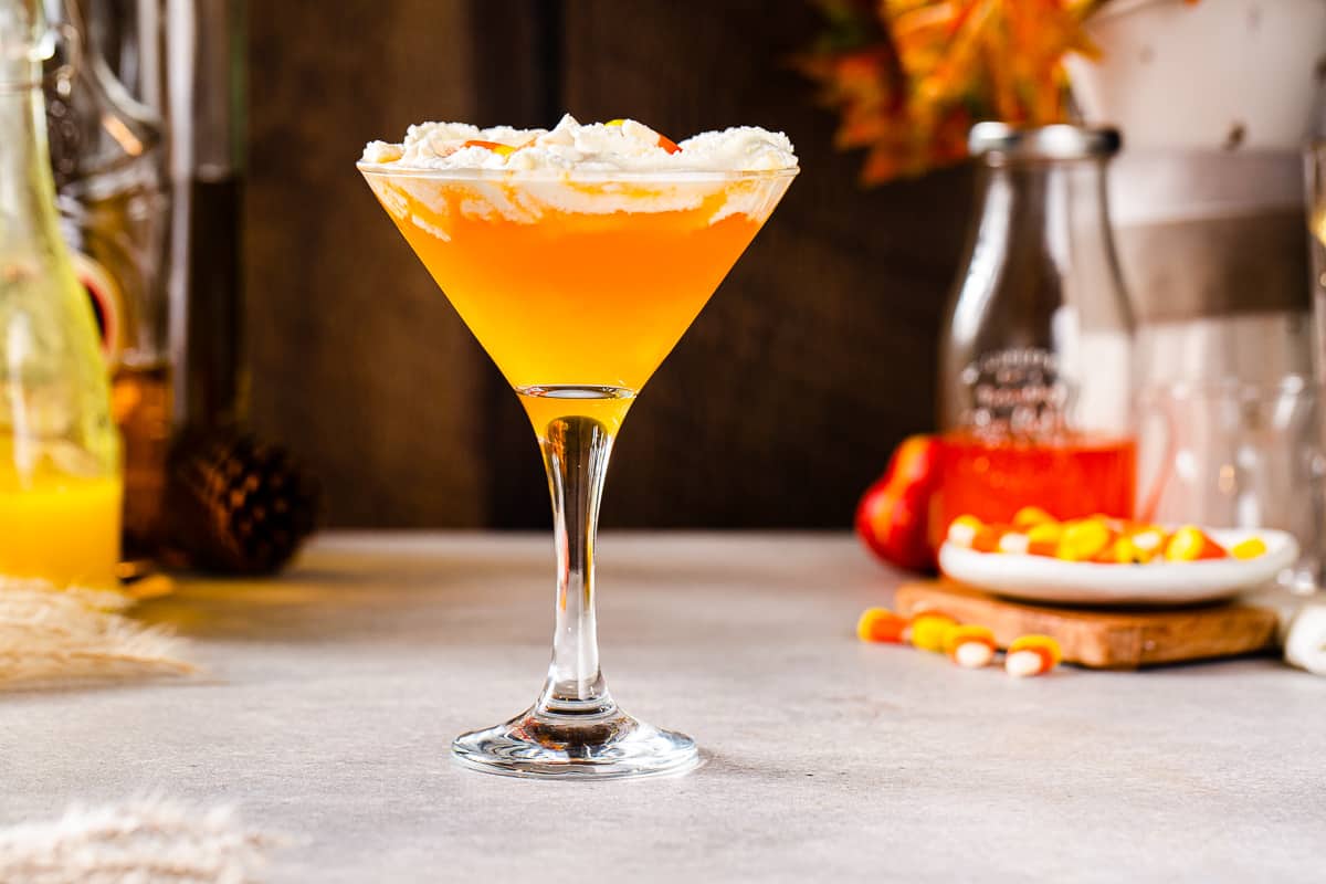 Side view of a Candy Corn Martini with three layers: the top layer is whipped cream, the middle layer is an orange liquid and the bottom layer is a yellow liquid. In the background is a plate of candy corn, bottles of ingredients and a container with orange autumn leaves.
