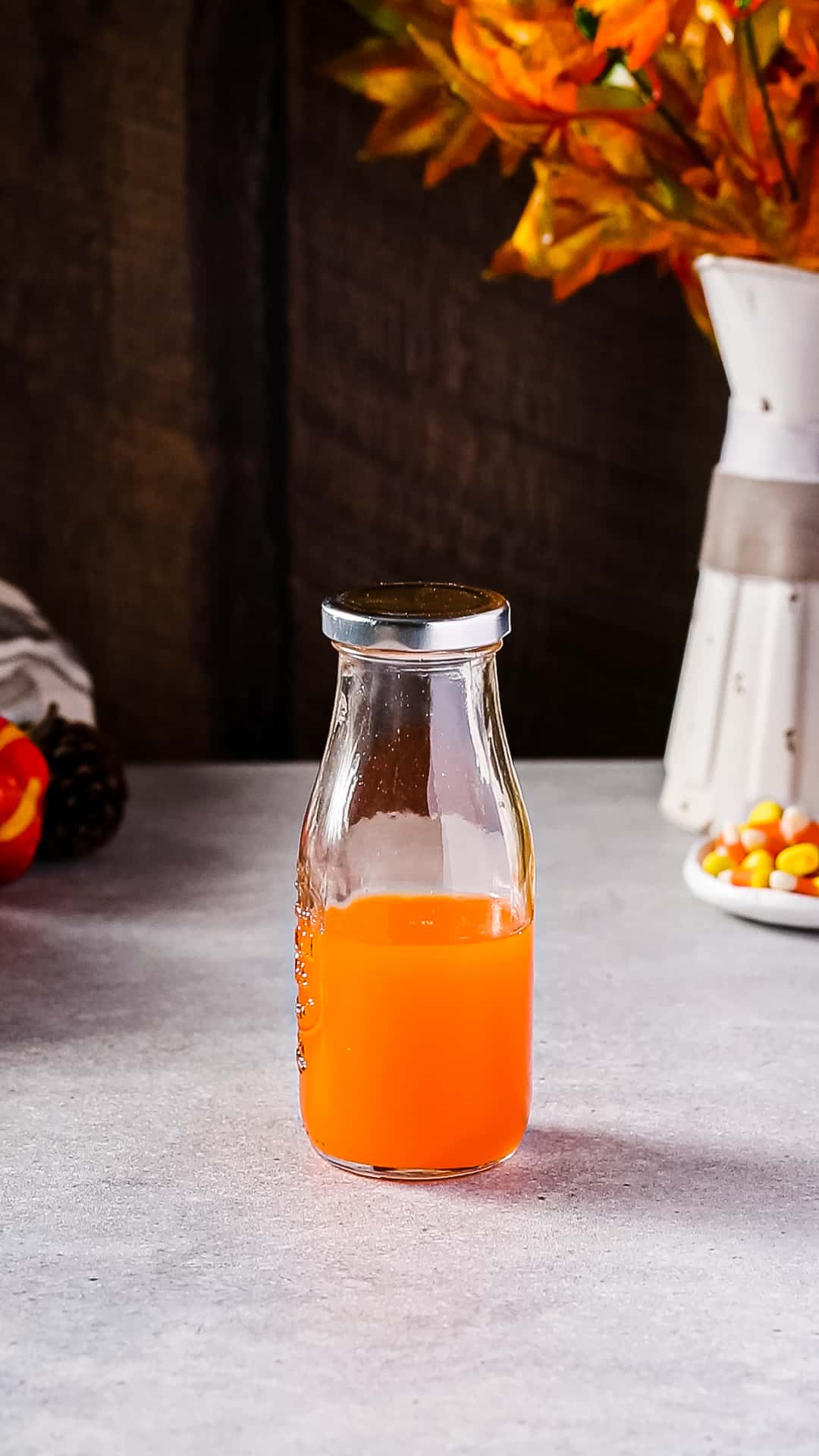 Bottle filled with orange liquid and capped with a silver cap on a tabletop. Candy corn and autumn leaves are in the background.