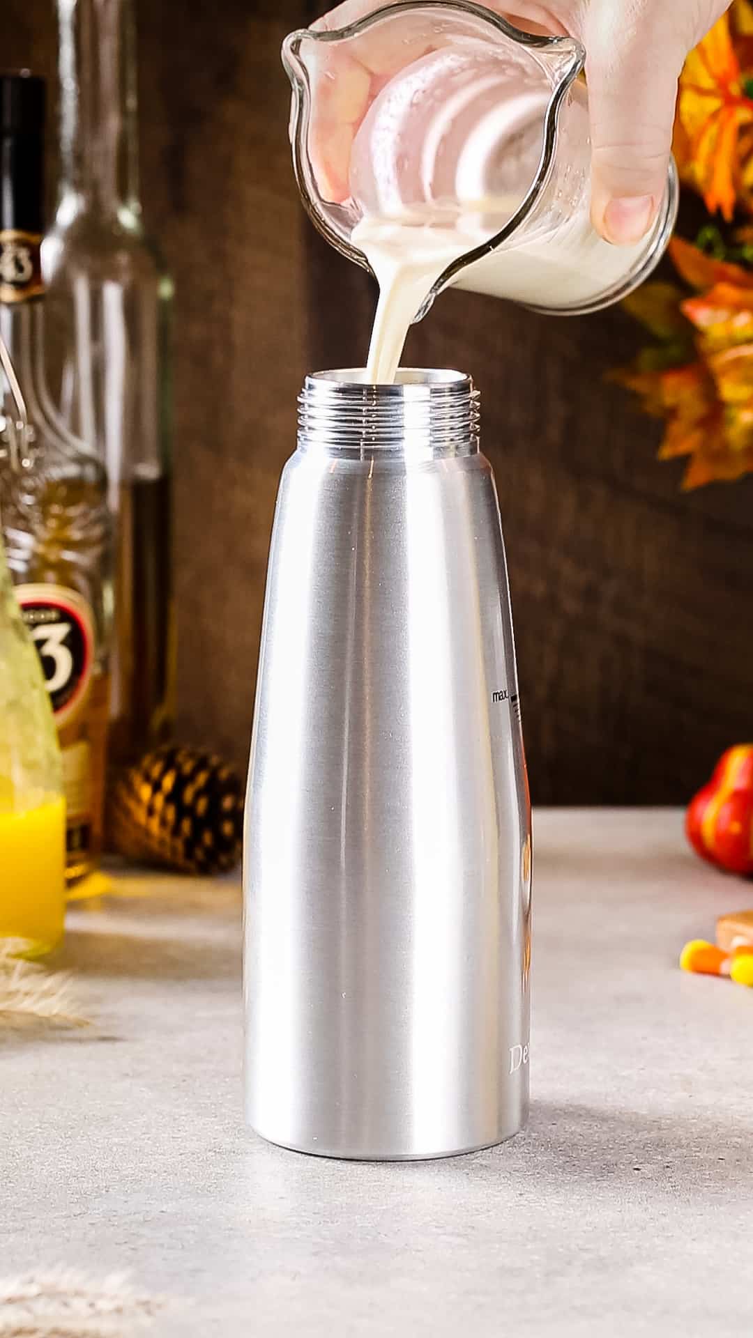 Hand pouring cream from a glass cup into a silver whipped cream dispenser.