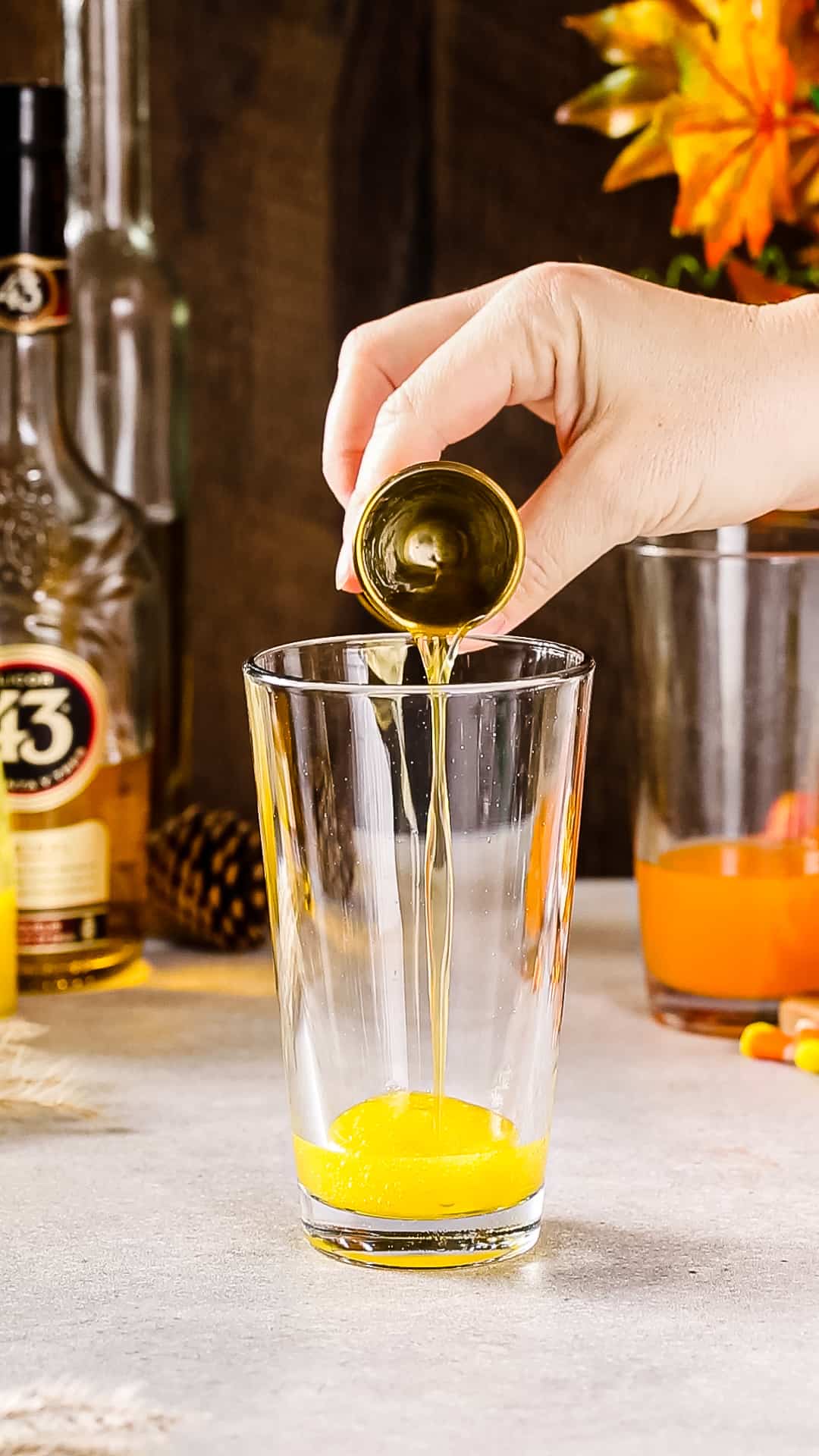 Hand pouring Licor 43 from a cocktail jigger into a cocktail shaker with yellow liquid in it.
