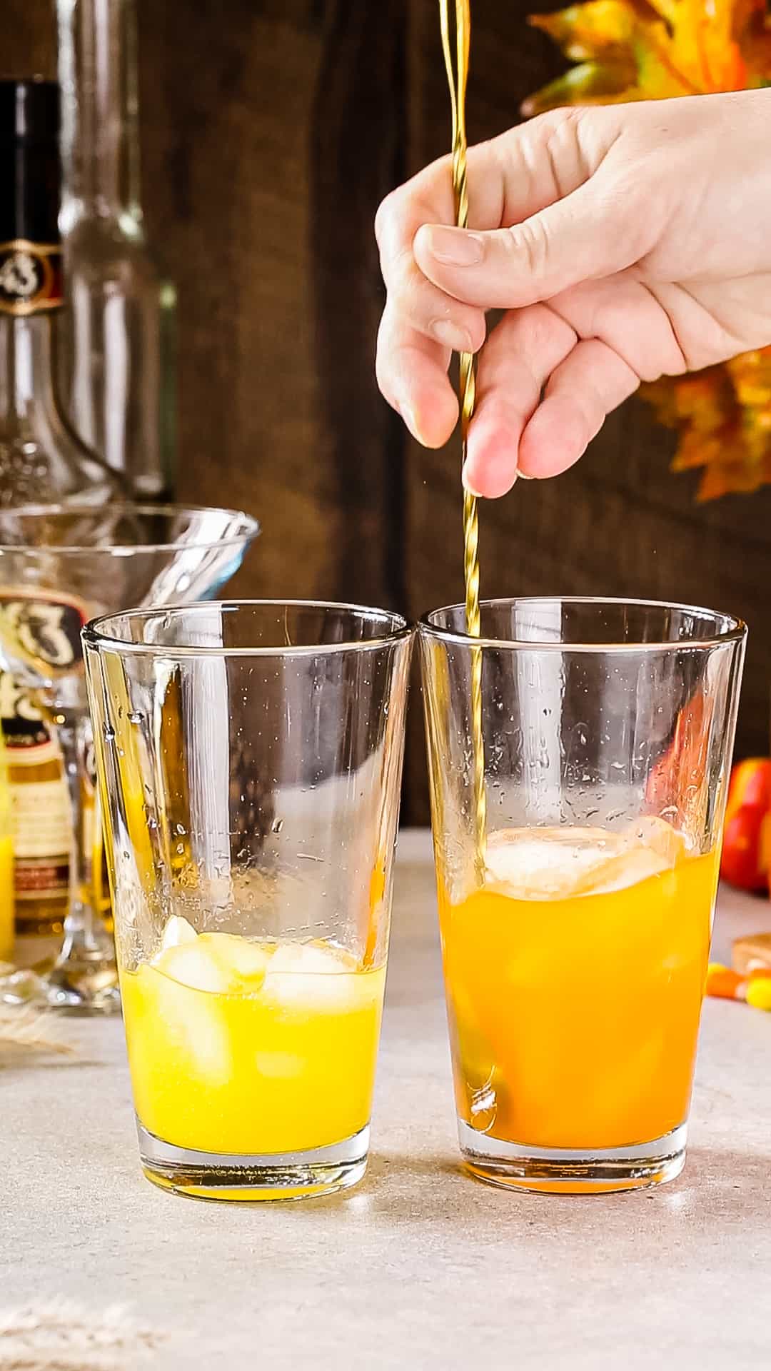Two glass cocktail shakers on a countertop, one with yellow liquid and ice, and one with orange liquid and ice. Hand is using a long bar spoon to stir the orange mixture.