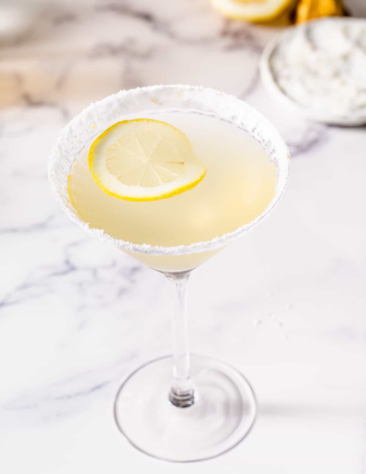 Overhead view of a Limoncello Martini with a sugared rim on a marble countertop. A cut lemon and a plate of sugar are in the background. A lemon slice is floating in the drink.