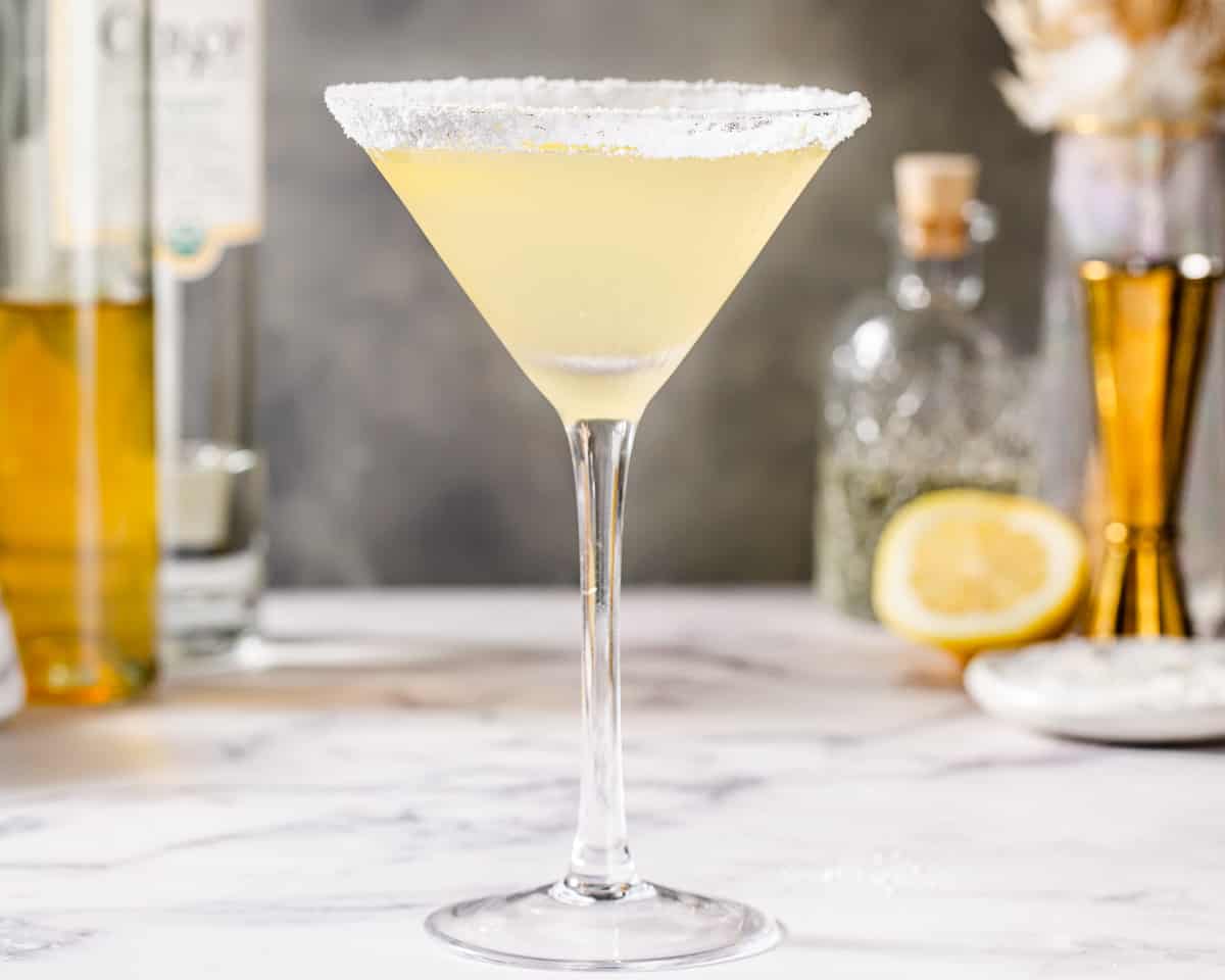 Side view of a yellow drink in a stemmed martini glass on a marble countertop. The rim is covered in sugar. Ingredients are in the background along with a gold jigger and dried flowers.