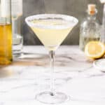 Side view of a yellow drink in a stemmed martini glass on a marble countertop. The rim is covered in sugar. Ingredients are in the background.
