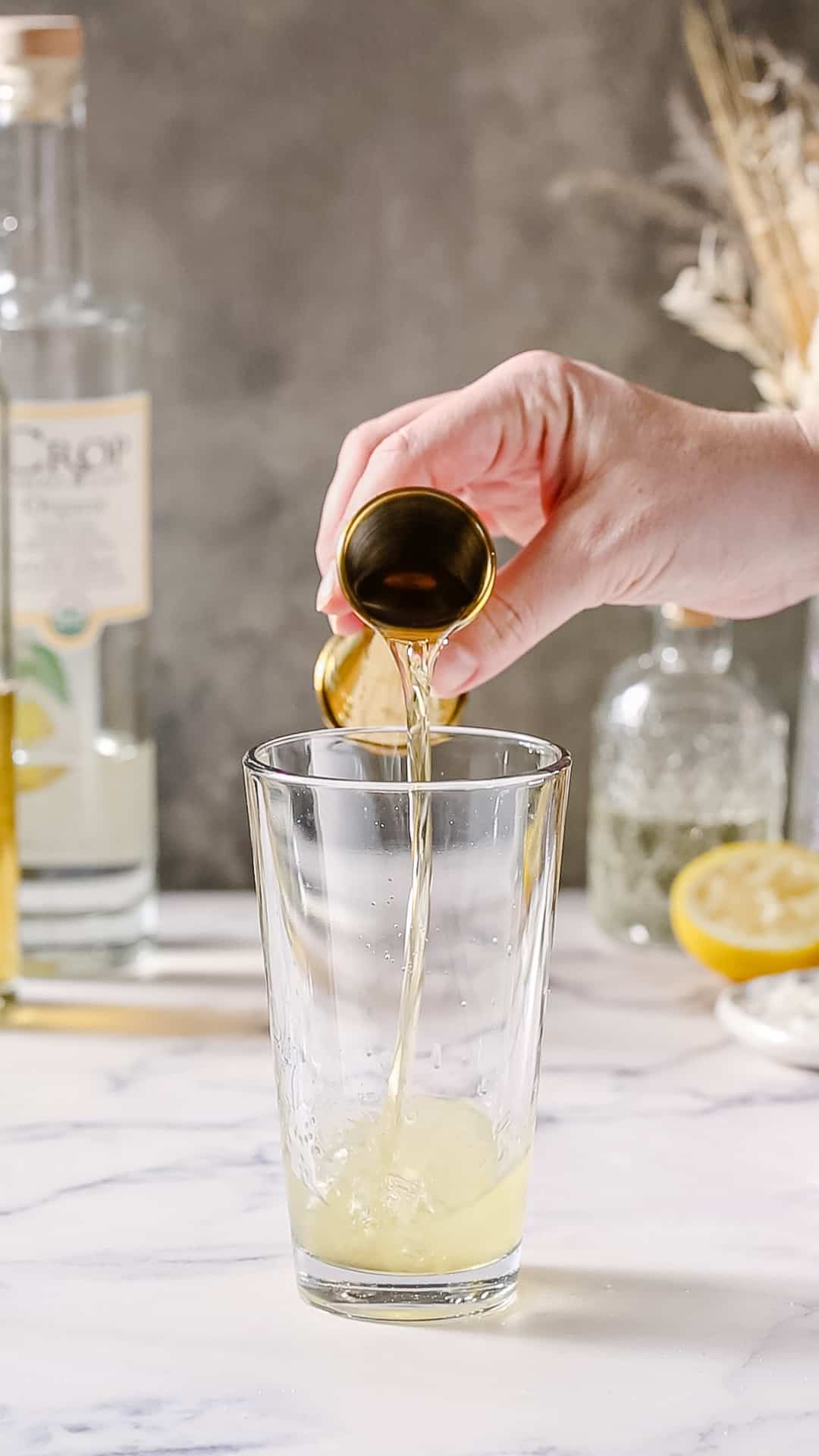 Hand using a gold jigger to pour limoncello liqueur into a cocktail shaker.