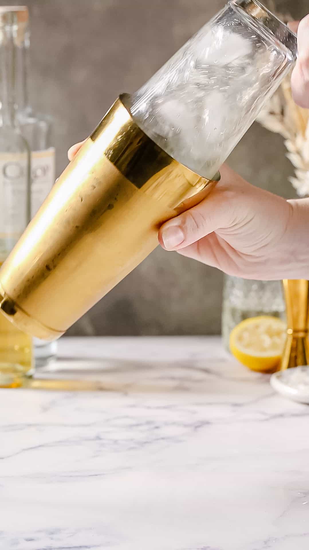 Hands shaking a liquid-filled cocktail shaker that has one gold side and one glass side.