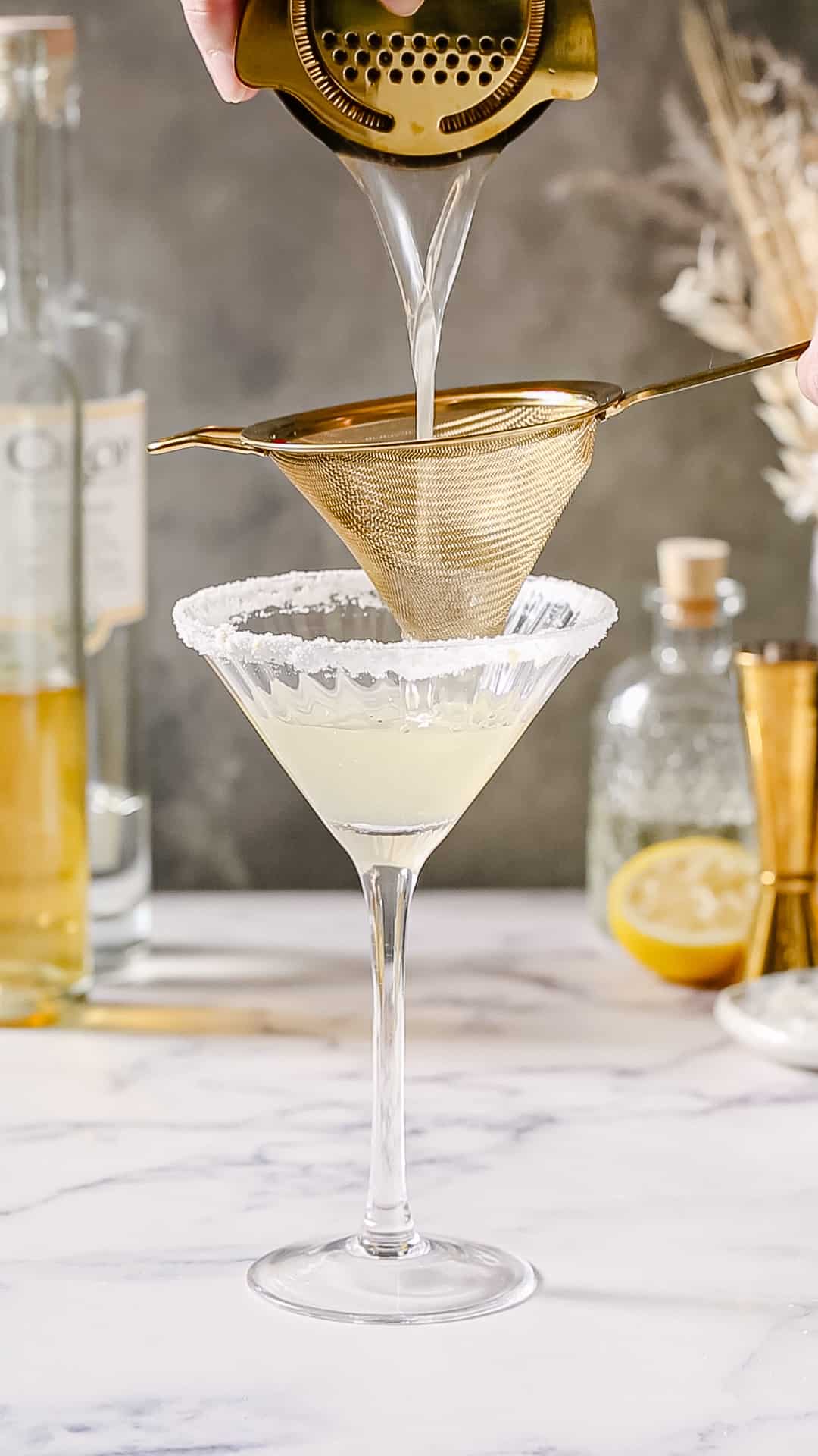 Hands using two strainers to strain a cocktail into a martini glass with a sugared rim.