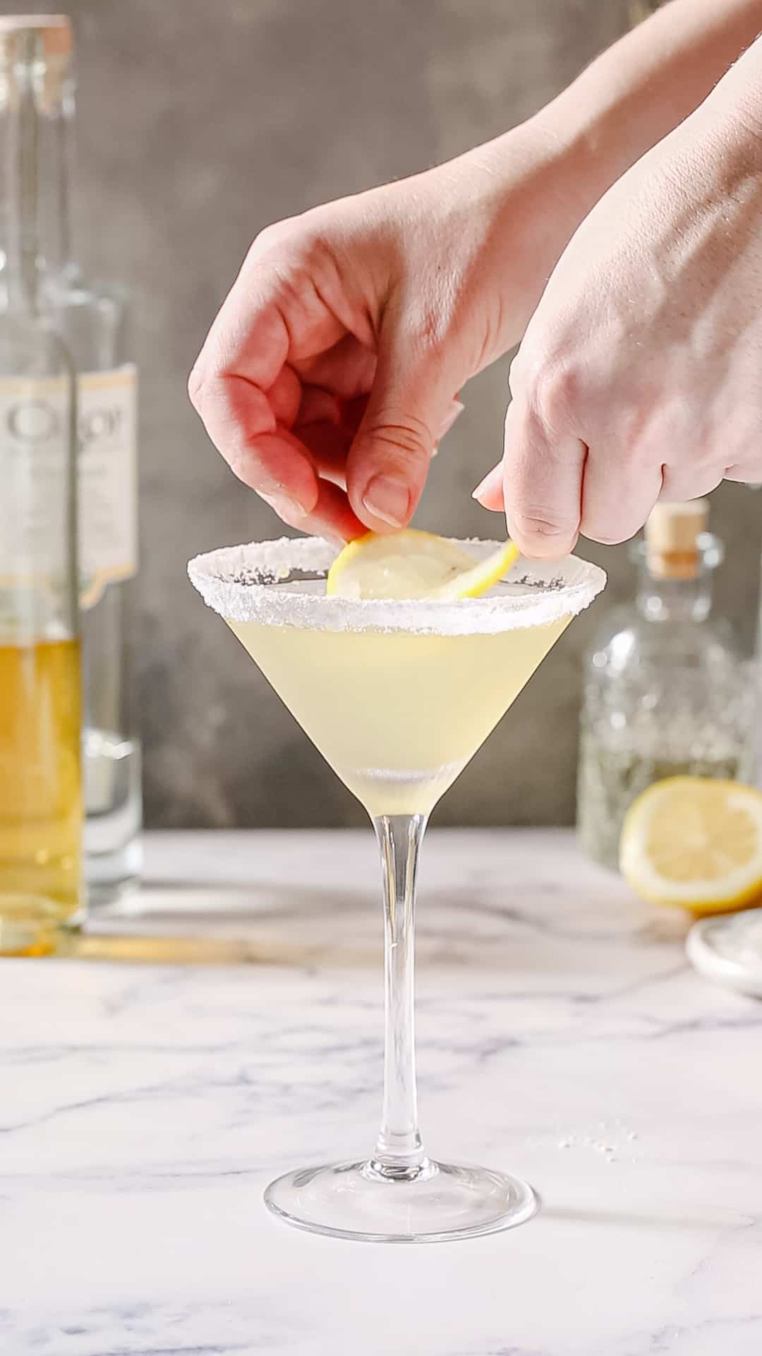 Hands adding a lemon slice to the surface of a yellow cocktail in a martini glass with a sugared rim.