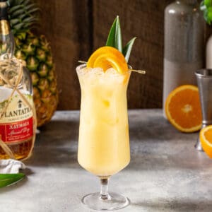 Side view of a yellow colored Painkiller cocktail in a Hurricane glass on a gray countertop with pineapple fronds and an orange slice garnish. In the background are cut oranges, pineapple and a bottle of rum.