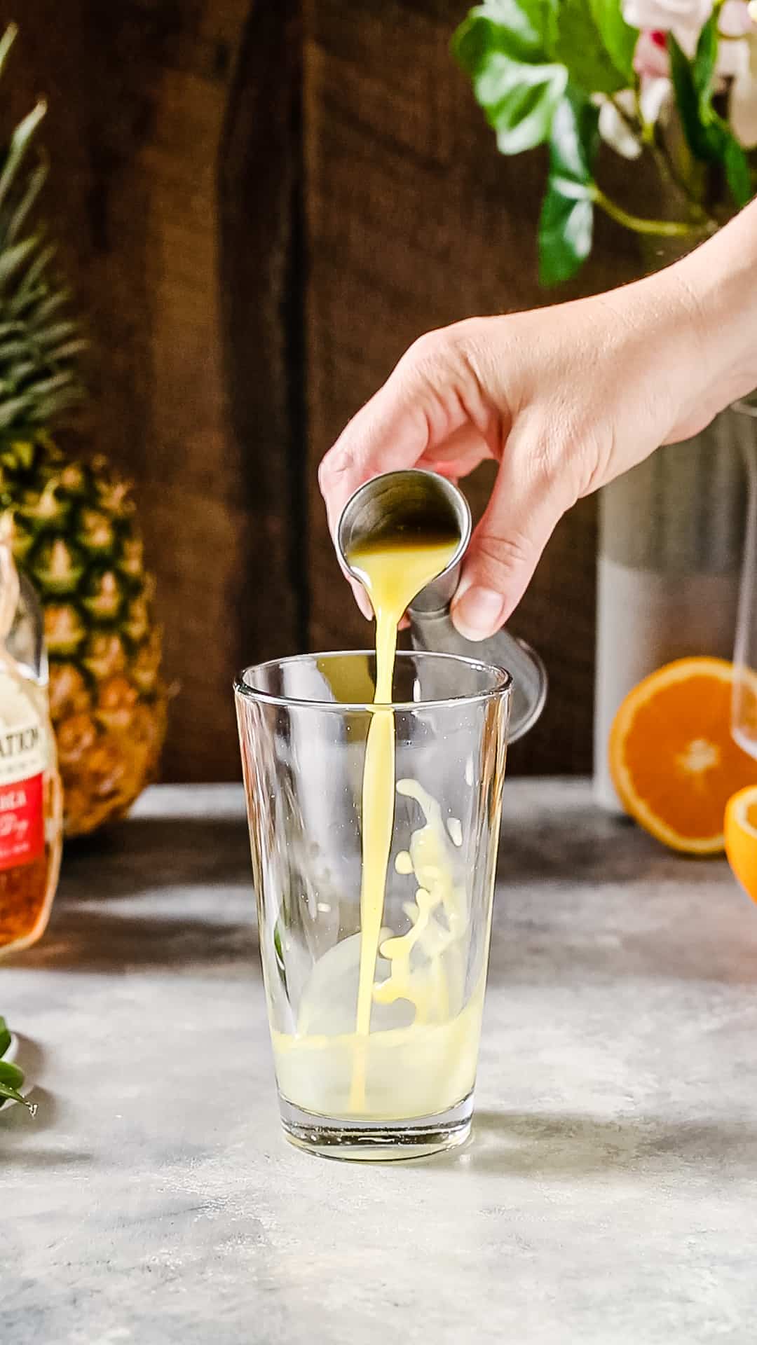 Hand pouring orange juice from a jigger into a cocktail shaker.