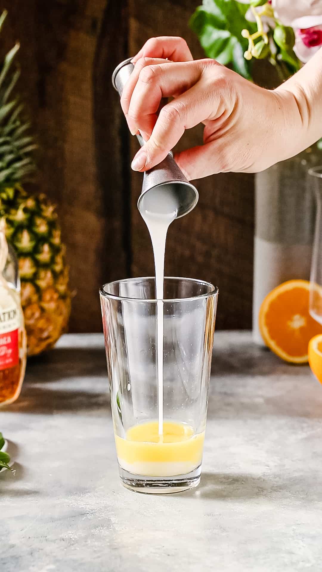 Hand pouring cream of coconut from a jigger into a cocktail shaker that is filled with orange juice.