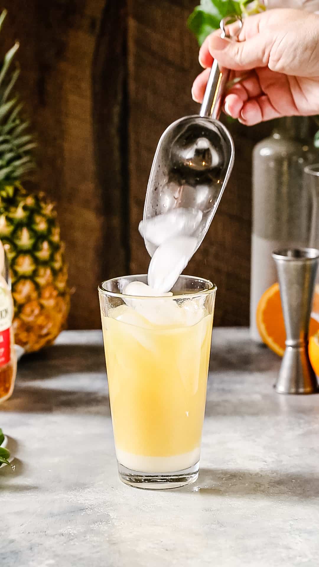 Hand using ice scoop to add ice to a glass cocktail shaker filled with yellow liquid.