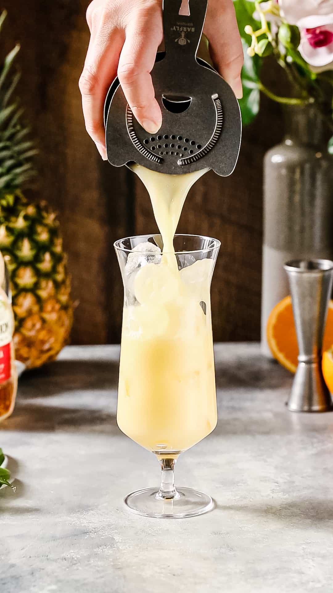 Hand using a gray cocktail strainer to strain the yellow drink into a hurricane glass filled with ice.