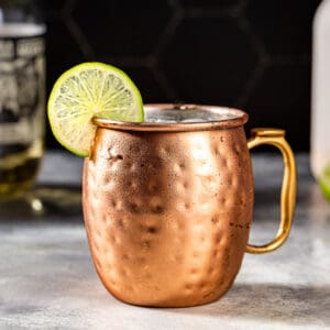 Side view of a Tequila Mule cocktail in a hammered copper mug. A lime wheel is on the rim of the mug as a garnish. In the background is a bottle of tequila.