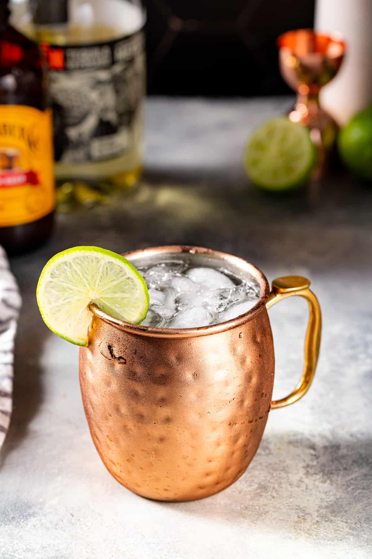 Overhead view of a Tequila Mule in a hammered copper mug. Crushed ice is visible in the mug and there is a lime wheel on the rim as a garnish. A bottle of ginger beer, a bottle of tequila, as well as a jigger and cut lime are in the background.