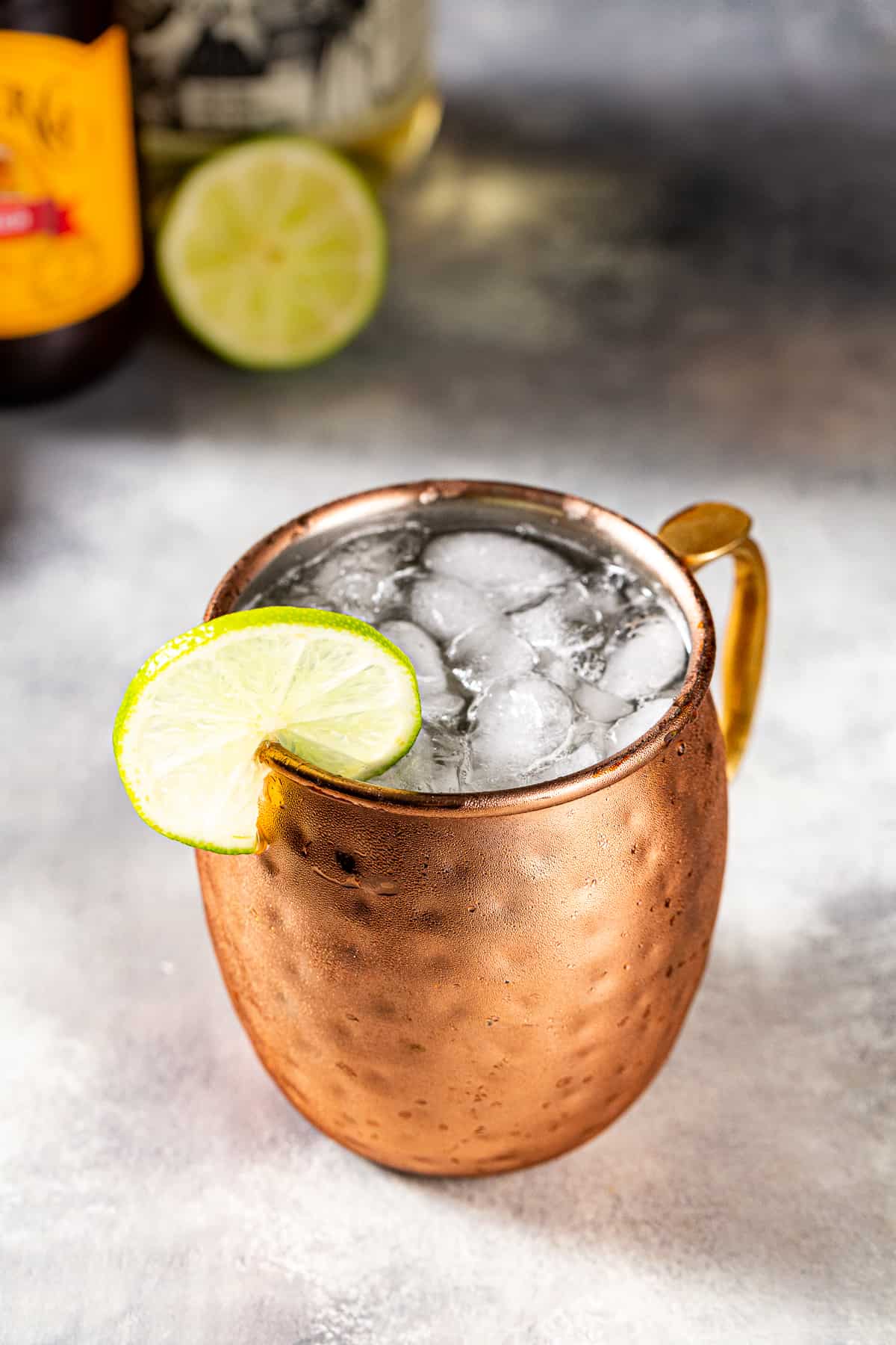 Overhead view of a Tequila Mule in a hammered copper mug. Crushed ice is visible in the mug and there is a lime wheel on the rim as a garnish. A bottle of ginger beer and a cut lime are in the background.