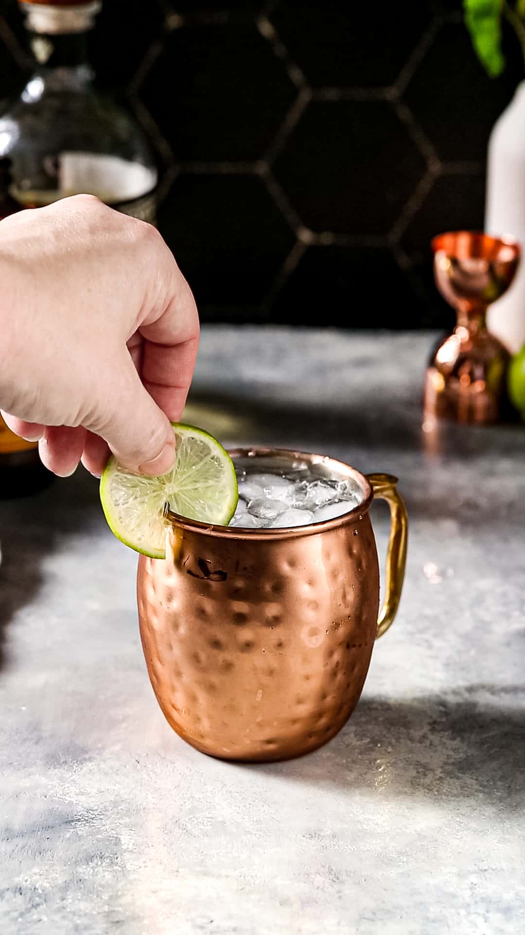 Hand adding a fresh lime wheel as a garnish to the rim of a copper mug filled with liquid and crushed ice.