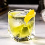 Side view of a White Negroni cocktail, which is a yellow colored cocktail in an old fashioned glass. The cocktail, which is on a marble countertop, is garnished with a twist of lemon, and there is a large piece of ice in the glass.