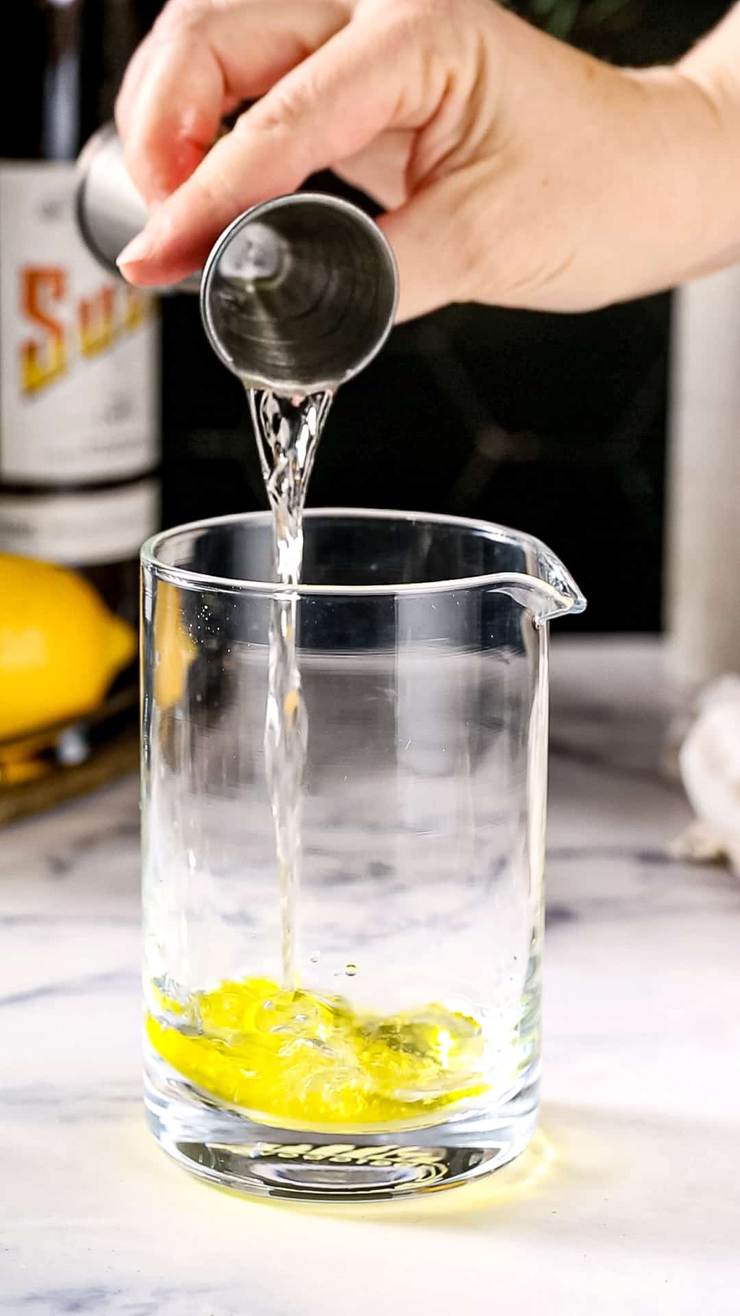 Hand using a jigger to pour dry vermouth into a mixing glass filled with yellow liquid.