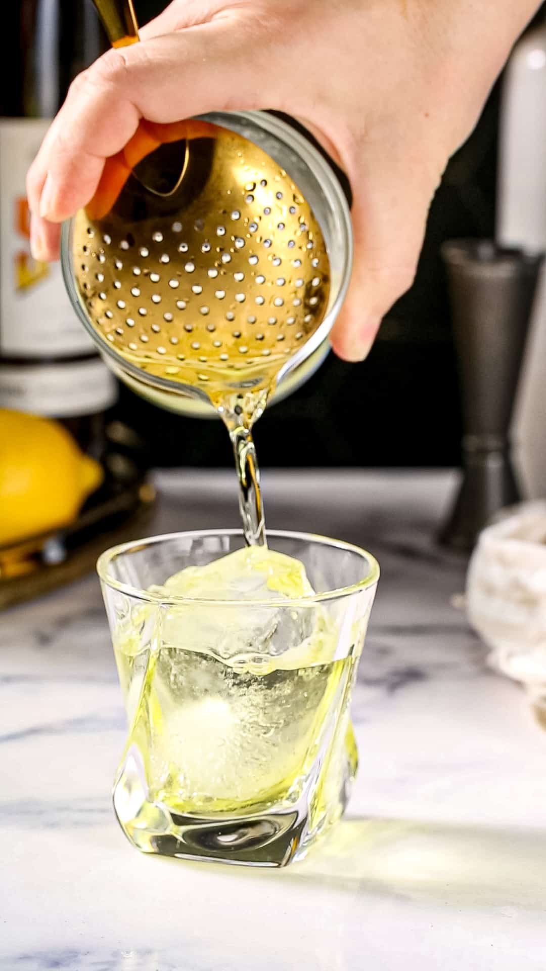 Hand using a Julep strainer to strain a yellow colored cocktail into the serving glass.