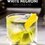 Side view of a White Negroni cocktail, which is a yellow colored cocktail in an old fashioned glass. The cocktail, which is on a marble countertop, is garnished with a twist of lemon, and there is a large piece of ice in the glass. Above the drink, text reads White Negroni gin cocktail.