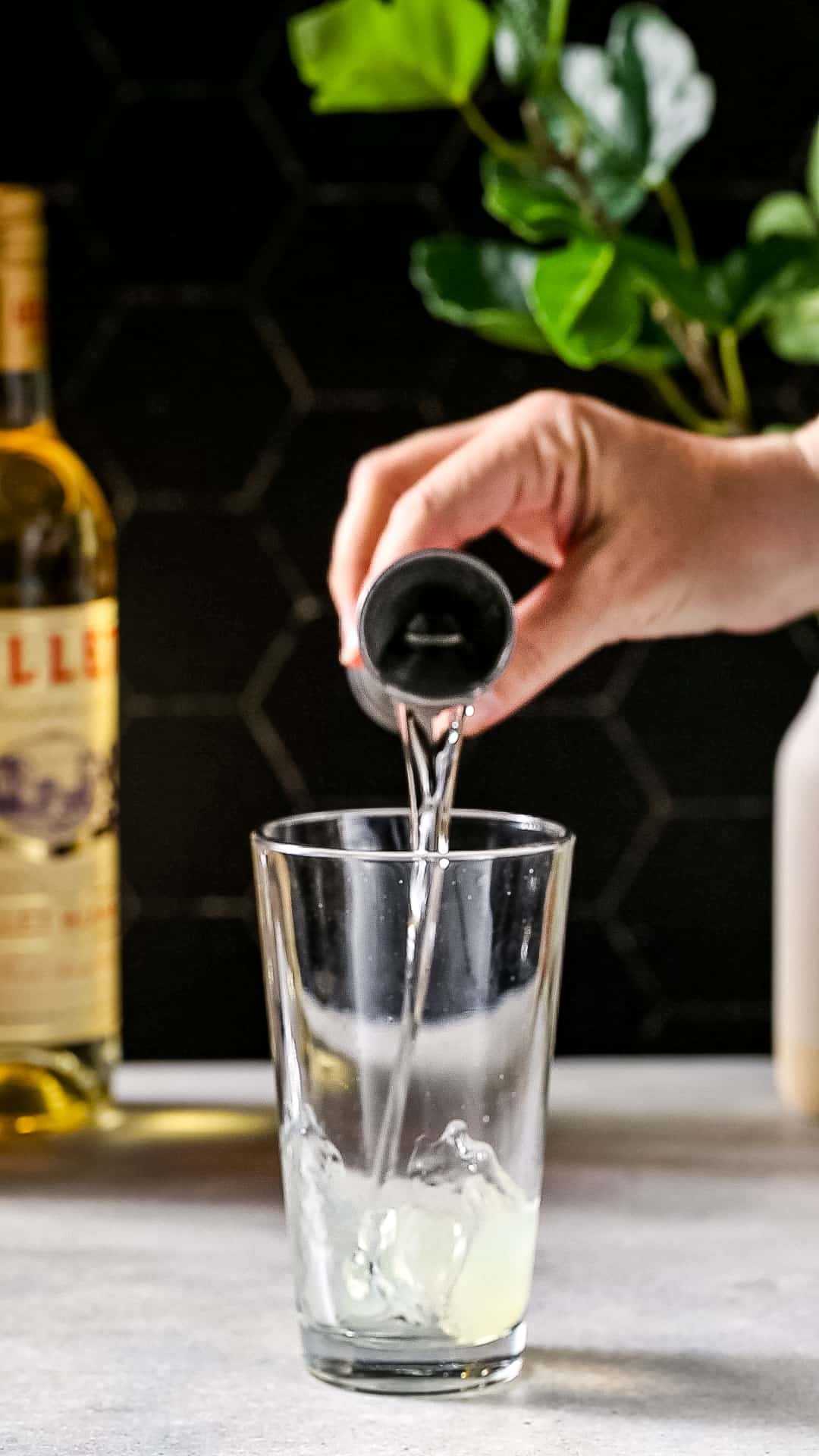Hand adding gin from a jigger into a cocktail shaker.