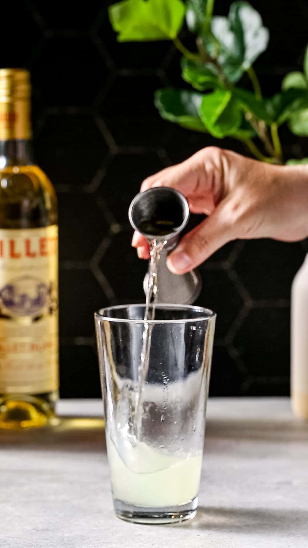 Hand adding Lillet Blanc from a jigger into a cocktail shaker.