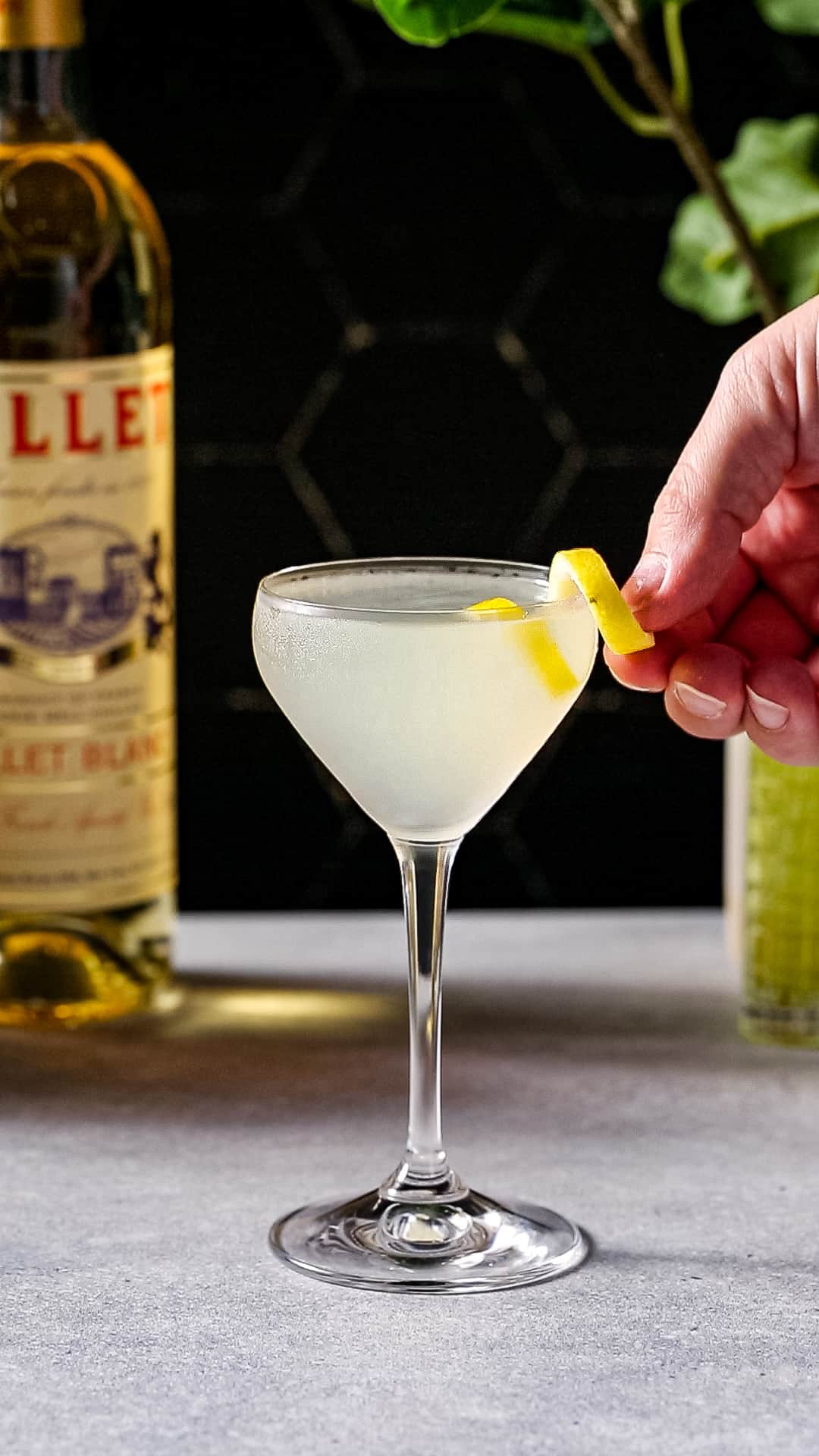 Hand adding a lemon peel garnish to a stemmed cocktail glass filled with yellow liquid.