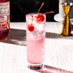 Dirty Shirley cocktail on a white wood countertop with maraschino cherries, grenadine and a gold colored jigger in the background.