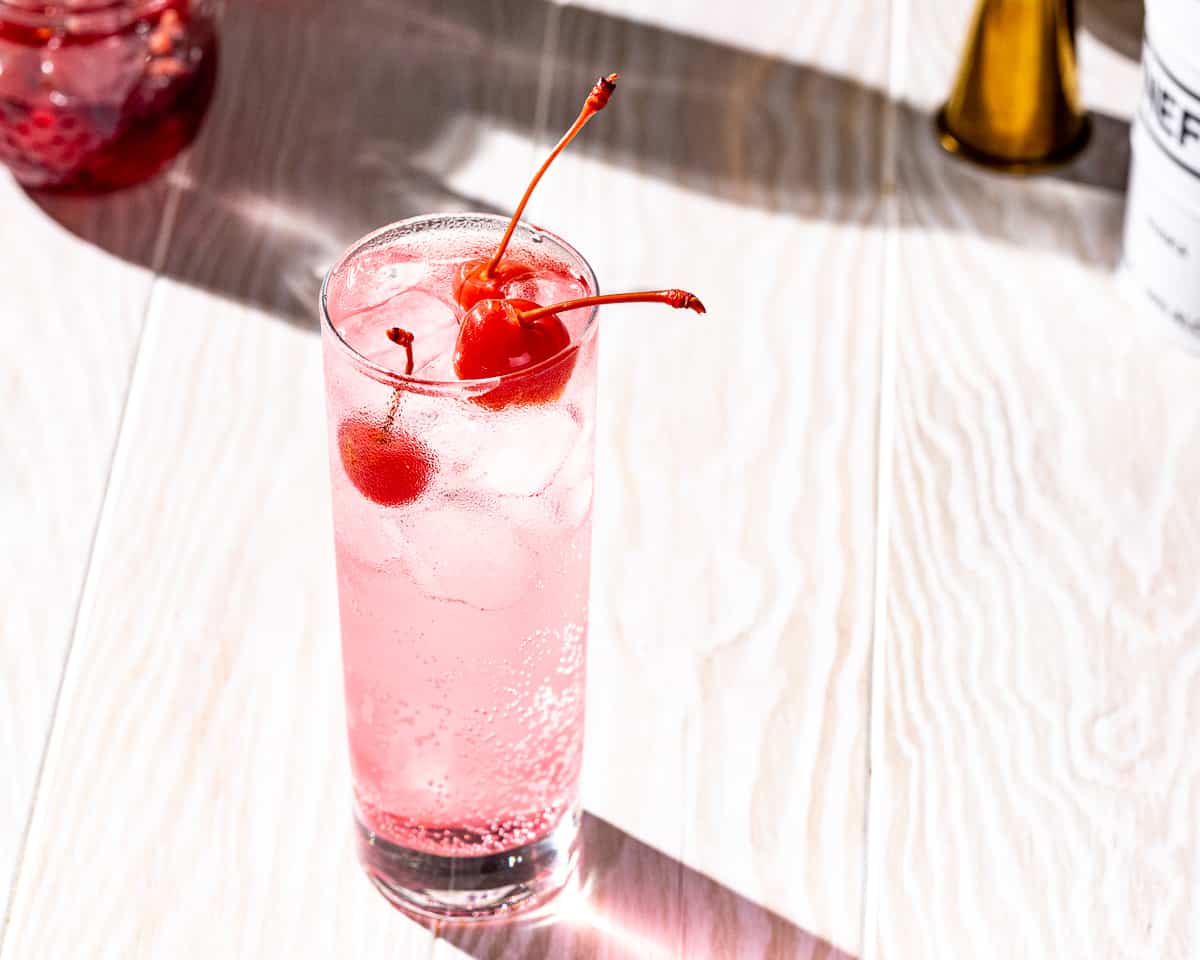 Overhead view of a pink colored Dirty Shirley cocktail with three maraschino cherries on a white wood countertop. In the background is a jar of maraschino cherries, a gold colored jigger and a container of vodka.